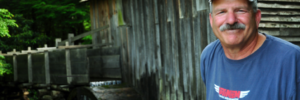 Man wearing a white baseball cap standing next to a wood shed