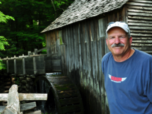 Man wearing a white baseball cap standing next to a wood shed